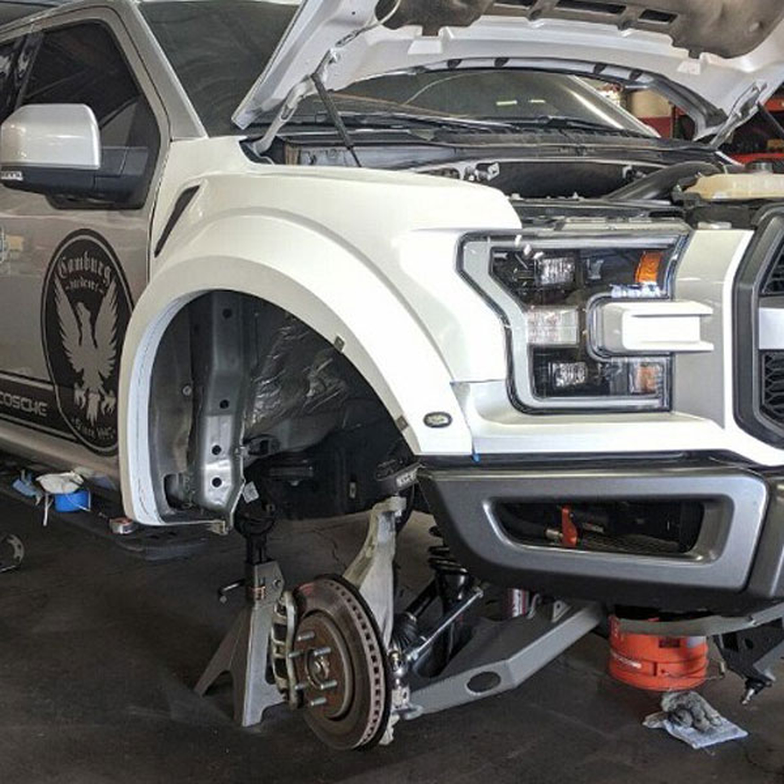 front end view of white raptor currently on jack stands with camburg long travel being installed