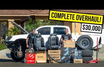 Two people stand in front of a white truck with its hood open, surrounded by various car parts and accessories like tires, seats, and boxes labeled Dirt King and Powertrax. A large yellow banner above them reads "Complete Overhaul $30,000."