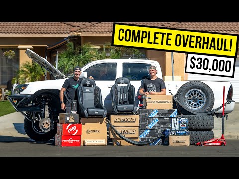 Two people stand in front of a white truck with its hood open, surrounded by various car parts and accessories like tires, seats, and boxes labeled Dirt King and Powertrax. A large yellow banner above them reads 