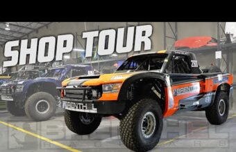 Off-road racing truck with orange and black livery parked inside a workshop with another similar truck in the background. Bold text above reads "Shop Tour".