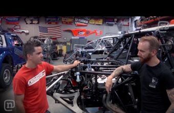 Two people are standing and talking in a garage with multiple off-road vehicles and equipment around them. One person is wearing a red shirt and the other is wearing a black shirt.