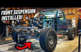 A teal modified truck with large off-road tires and no front body panels is in a garage. The vehicle has a custom front suspension and roll cage typical of a prerunner. A red arrow points to the suspension area. Tools and equipment are in the background.