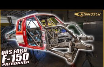 A partially assembled off-road vehicle frame with a roll cage and cab, specifically an OBS Ford F-150 prerunner, inside a workshop. The vehicle's front suspension and frame components are visible, and the KibbeTech logo is in the top right corner.