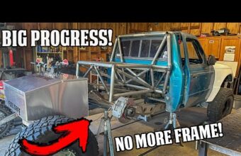 A blue truck in a garage is undergoing modifications. The rear frame is removed, showing a metal support structure. A fuel cell and large tire are visible along with tools and equipment. Text overlays read "Big Progress" and "No More Frame" with an arrow pointing at the tire.