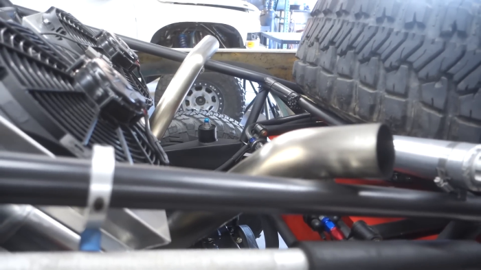 Close-up of a vehicle's engine compartment with visible components including pipes and fans, a large off-road tire is mounted inside the rear bed, another vehicle with similar tires in the background, possibly a prerunner in a garage setting.