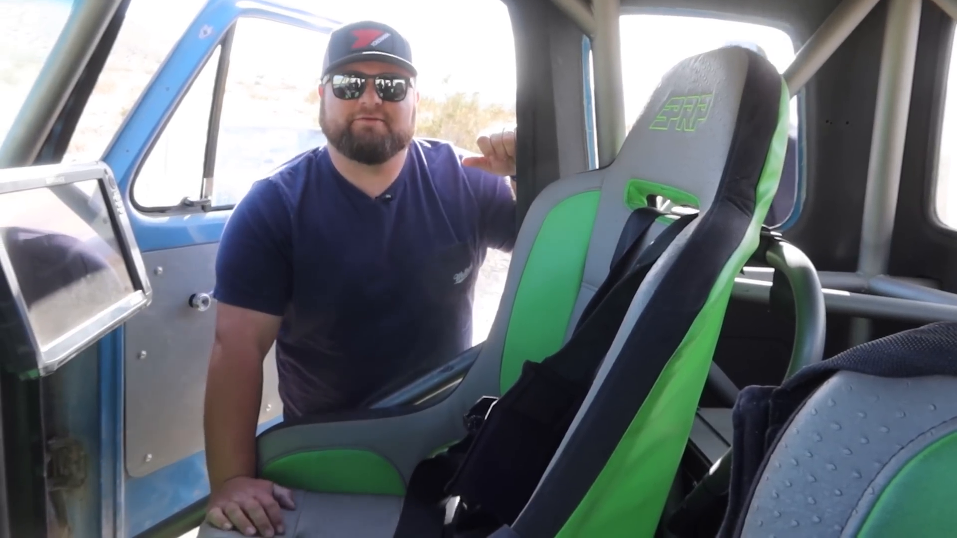 A man wearing sunglasses and a cap is inside a blue vehicle, standing beside a racing seat with green and gray upholstery. The vehicle has a minimalist interior design and visible roll cage, typical for a prerunner.