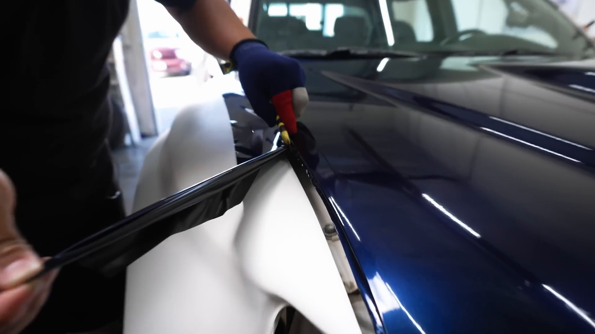 A person wearing a glove uses a tool to apply a wrap to the fender of a blue vehicle, likely a prerunner, in a garage setting.