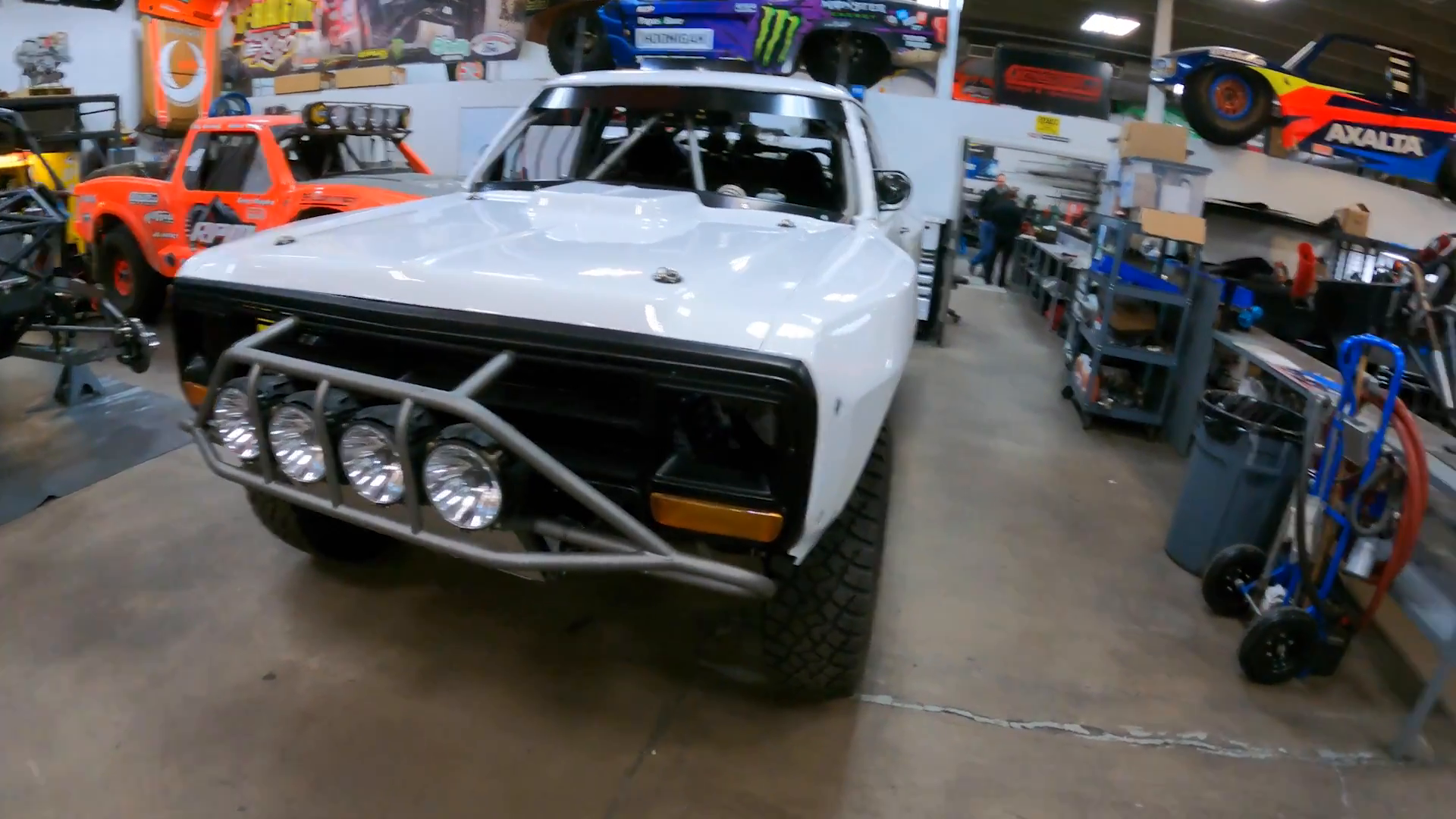 White prerunner truck with a front grille guard and off-road lights in a workshop, multiple off-road vehicles are on display, and there are tools and equipment on shelves.