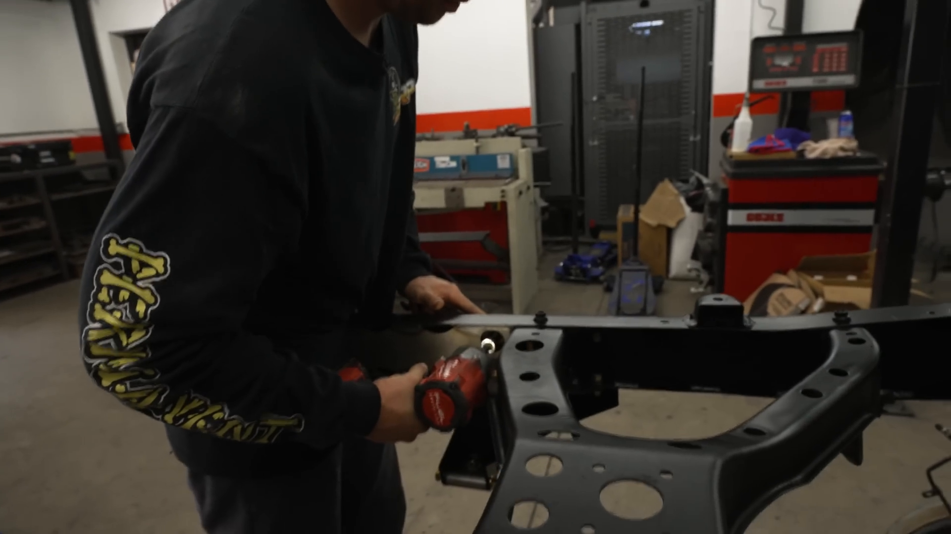 A person uses a power tool to work on a black metal vehicle frame in a workshop. Shelving units, automotive equipment, and tools are visible in the background.