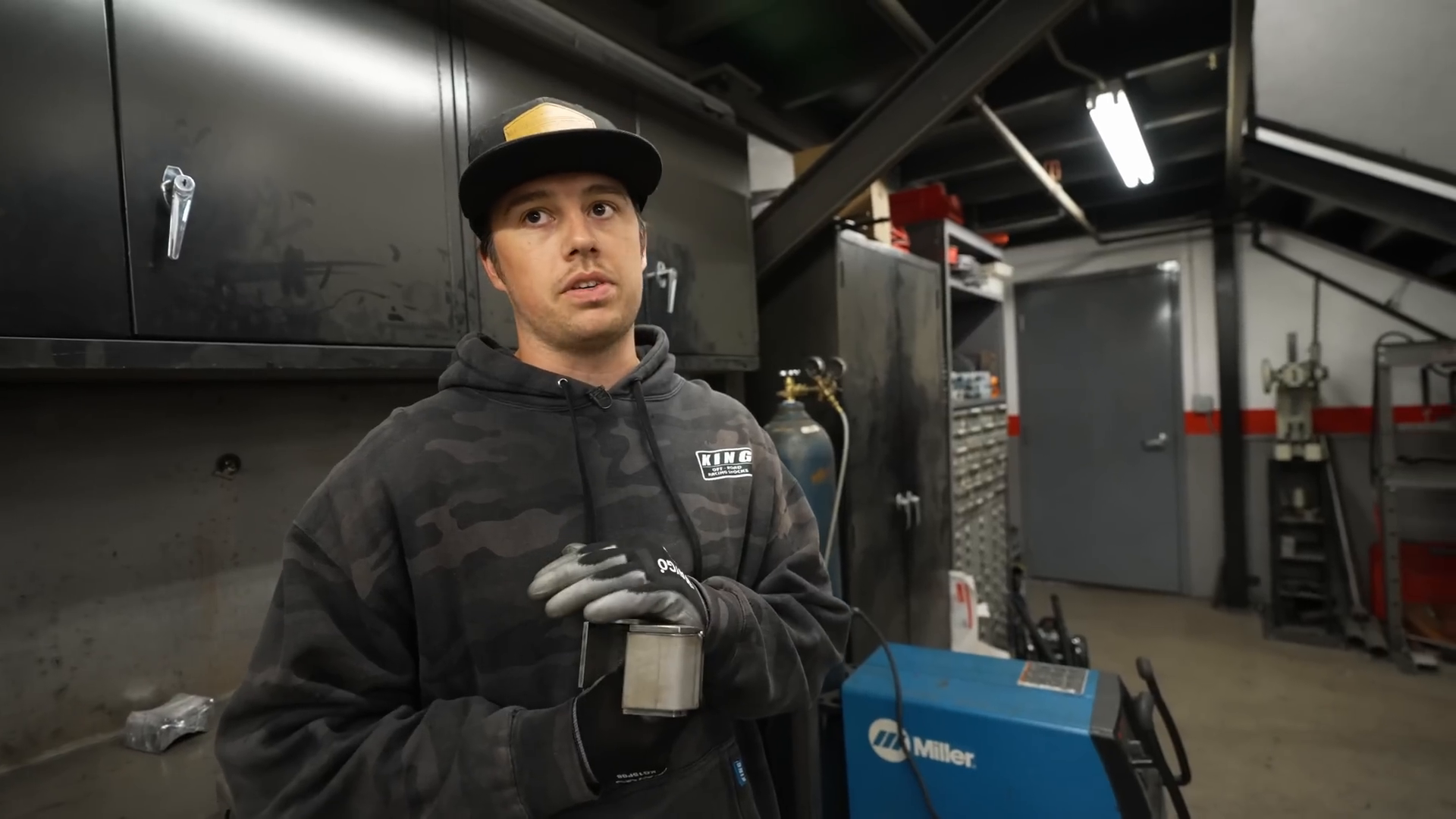 A person wearing a dark hoodie and cap stands in a garage or workshop. They are holding a metal component and wearing gloves. Behind them are black cabinets, a gas cylinder, and a welding machine. The space is organized with various tools and equipment.