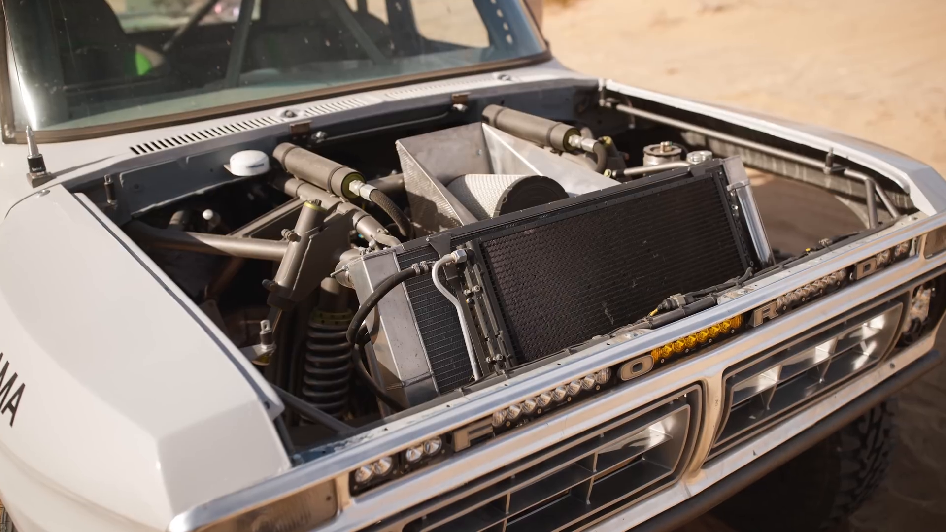 A modified vehicle's engine bay with exposed components including a radiator, suspension system with coilovers, and metal tubing, typical of a prerunner style truck.