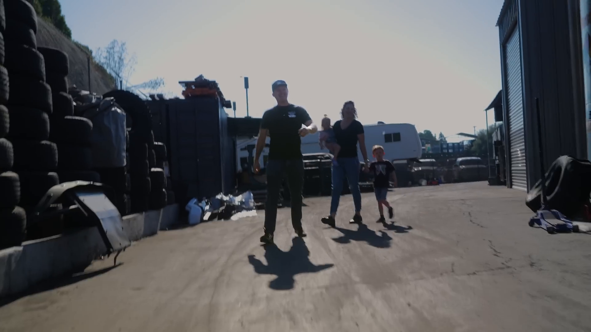 A group of people including a man, a woman holding a child, and another child are walking on a dirt path. In the background are stacks of tires and parked vehicles, including a camper trailer. The setting appears to be an industrial area.