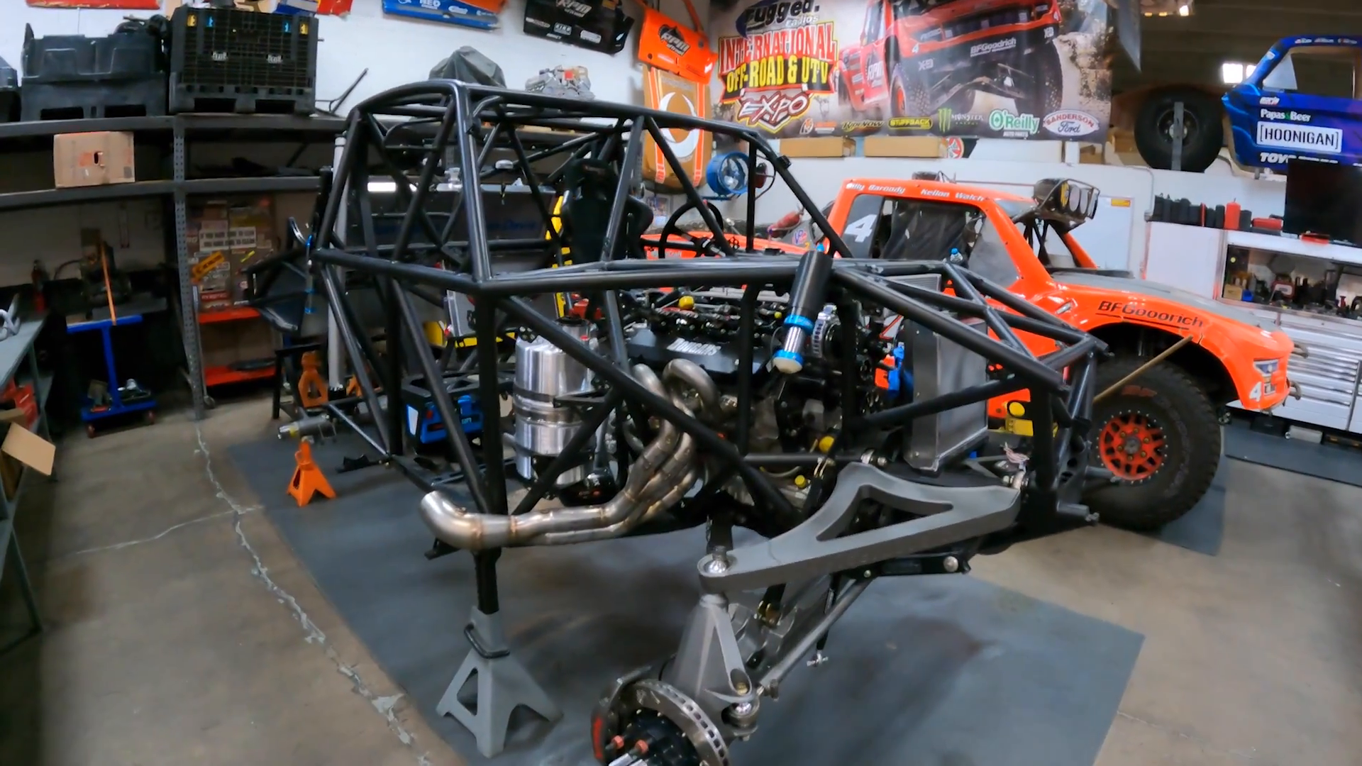 A garage with a partially built off-road vehicle frame on jack stands, an orange prerunner truck in the background, shelves with tools and equipment, and various posters on the walls.