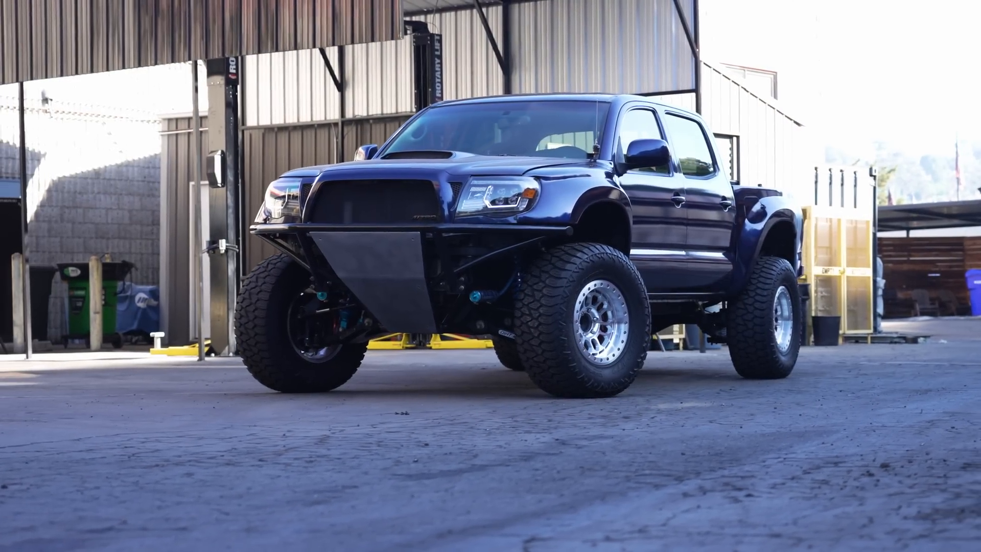 A modified pickup truck with large off-road tires and a custom front bumper parked in an industrial area.