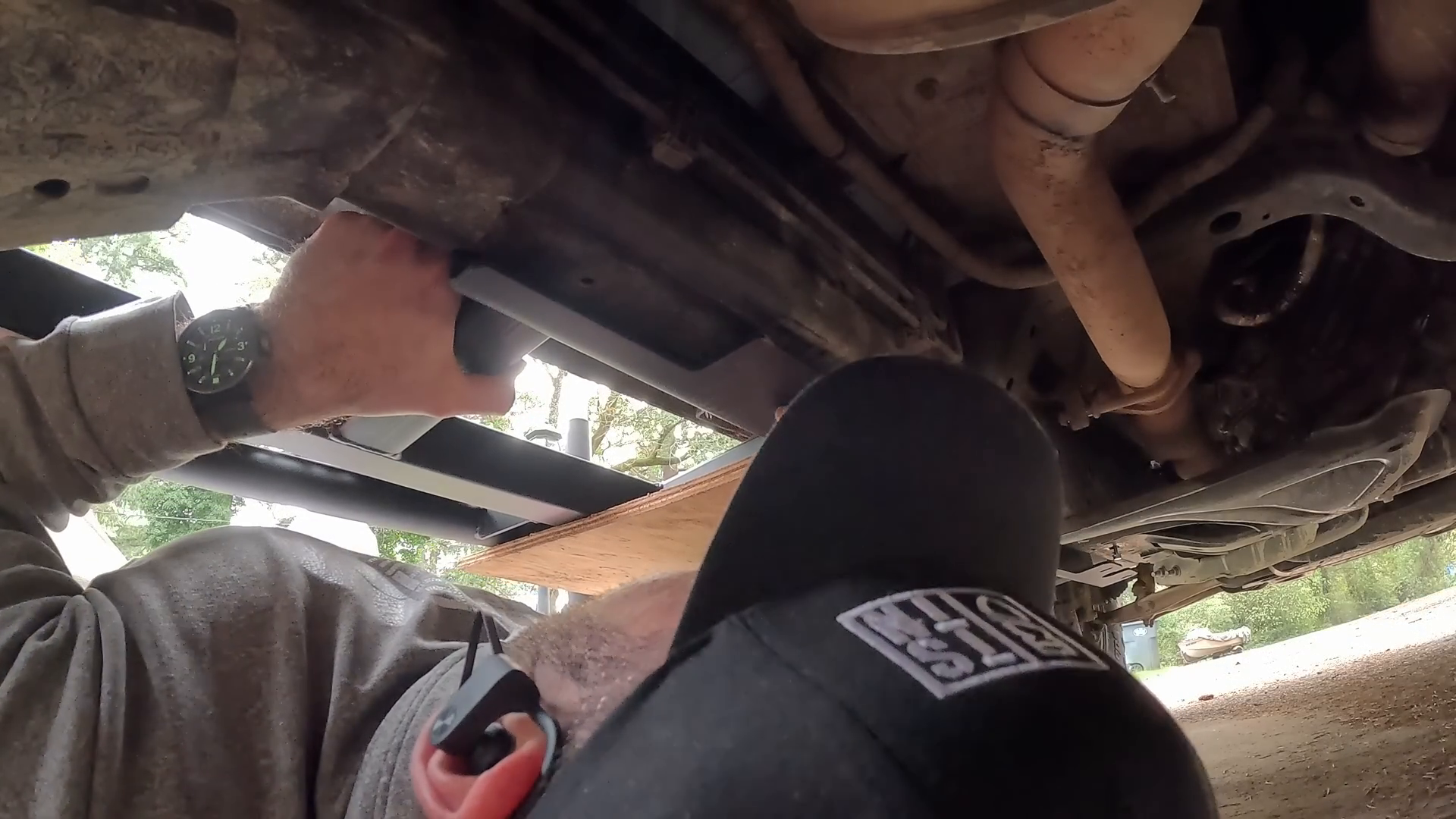 Person wearing a hat working underneath a vehicle, with one hand gripping a component. Visible parts include the exhaust pipe and chassis. Dirt and sunlight are visible, suggesting an outdoor setting.