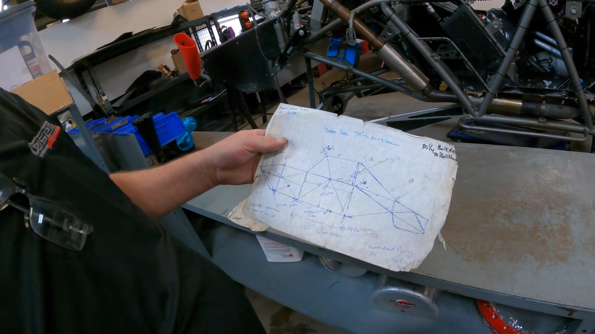 Person holding a hand-drawn diagram related to Prerunner design with measurements and notes in a workshop environment, visible are parts of a vehicle chassis and tools on a table.