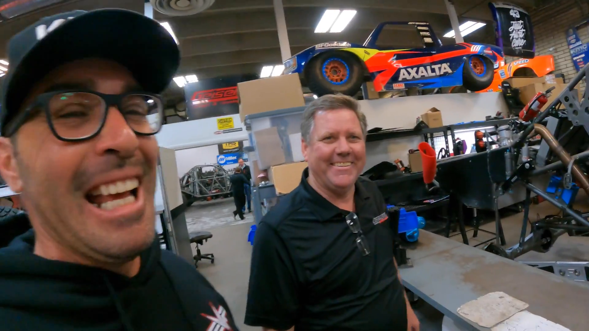 Two men standing in a workshop with vehicle parts and tools around them, a brightly-colored toy truck model painted with sponsor logos displayed on a shelf above.