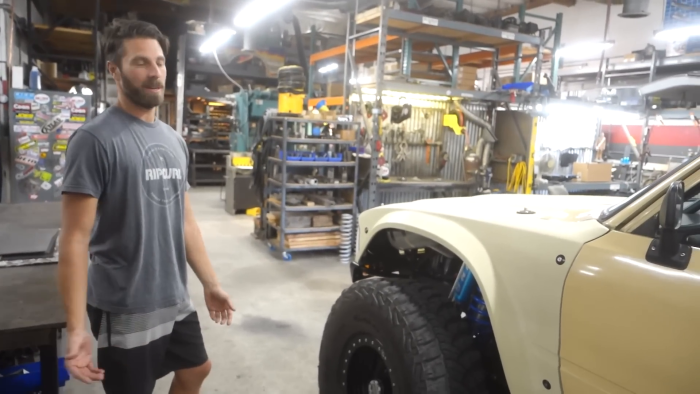 A person stands in a workshop next to a light-colored prerunner truck with large tires and visible suspension upgrades. The background shows tools, equipment, and shelves stocked with various items.