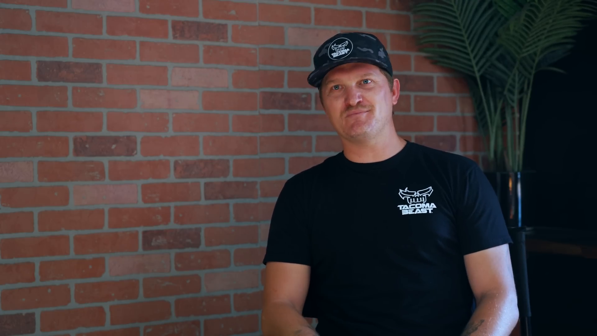 Man wearing a black Tacoma Beast t-shirt and a cap, sitting in front of a red brick wall with a potted plant in the background.