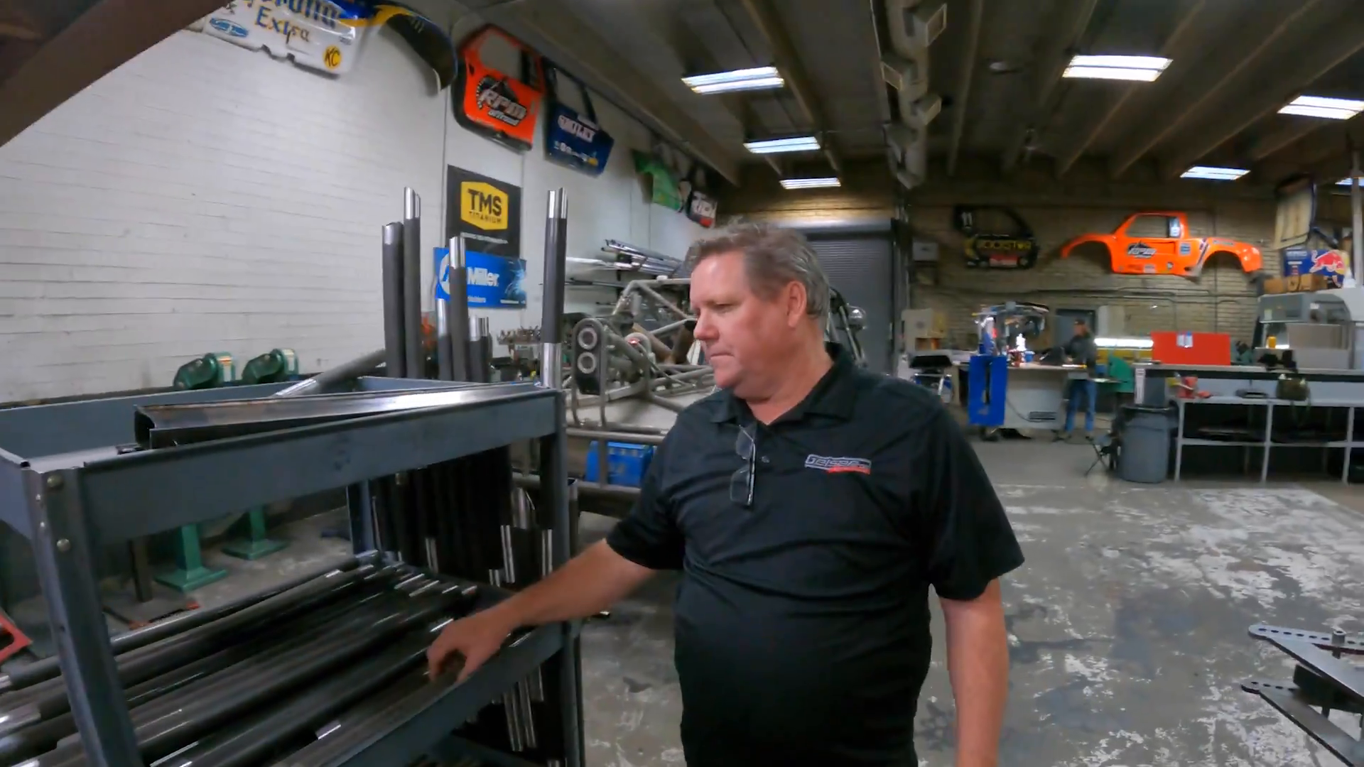 A man wearing a black shirt stands near metal pipes on shelves in a workshop. The workshop contains machinery, car parts, and a Prerunner body mounted on the wall. Various signs and banners are displayed on the walls.