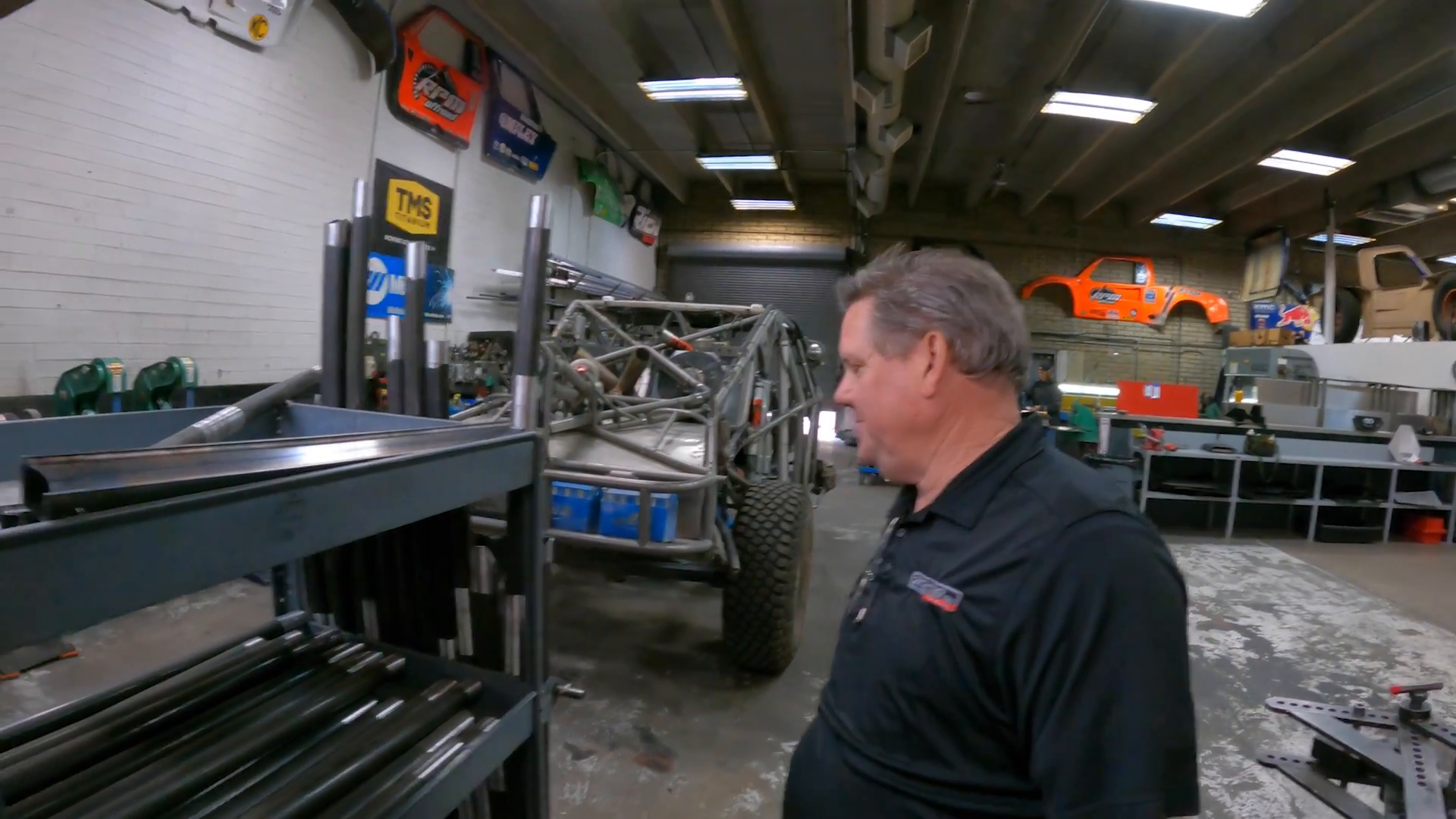 Man in a workshop with a vehicle chassis on large off-road tires in the background, various tools and equipment around, and several wall-mounted Prerunner truck models.