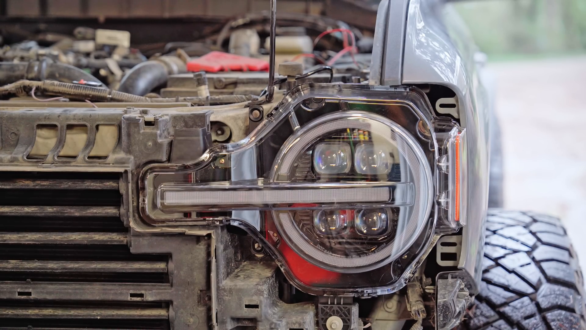 Front of a vehicle with a focus on the round LED headlight, grill, and exposed internal components. Visible rugged off-road tire, likely of a Prerunner, with wires and hoses around the engine area.