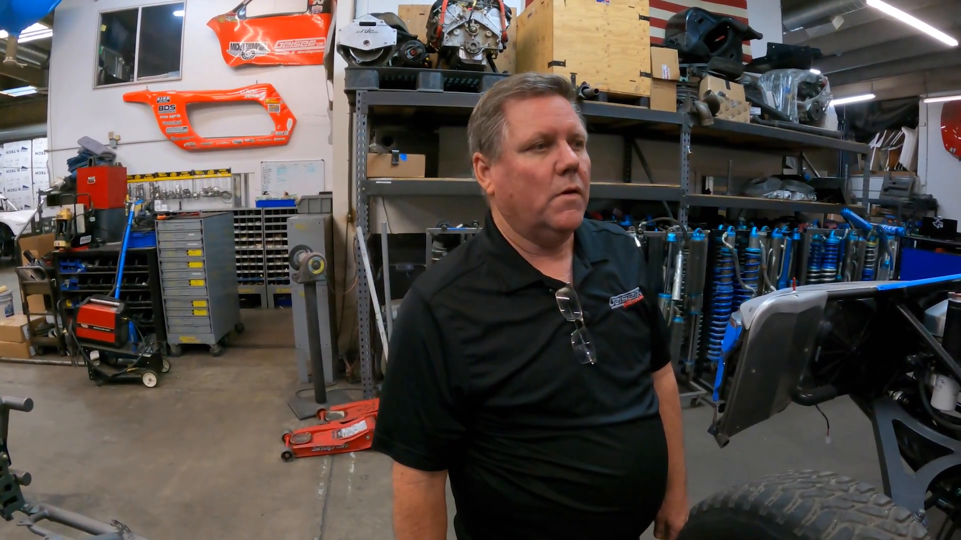 A man in a black polo shirt stands in a workshop with various tools and equipment including shocks, metal cabinets, an orange motorcycle part on the wall, and a partially visible off-road vehicle or prerunner in the foreground.