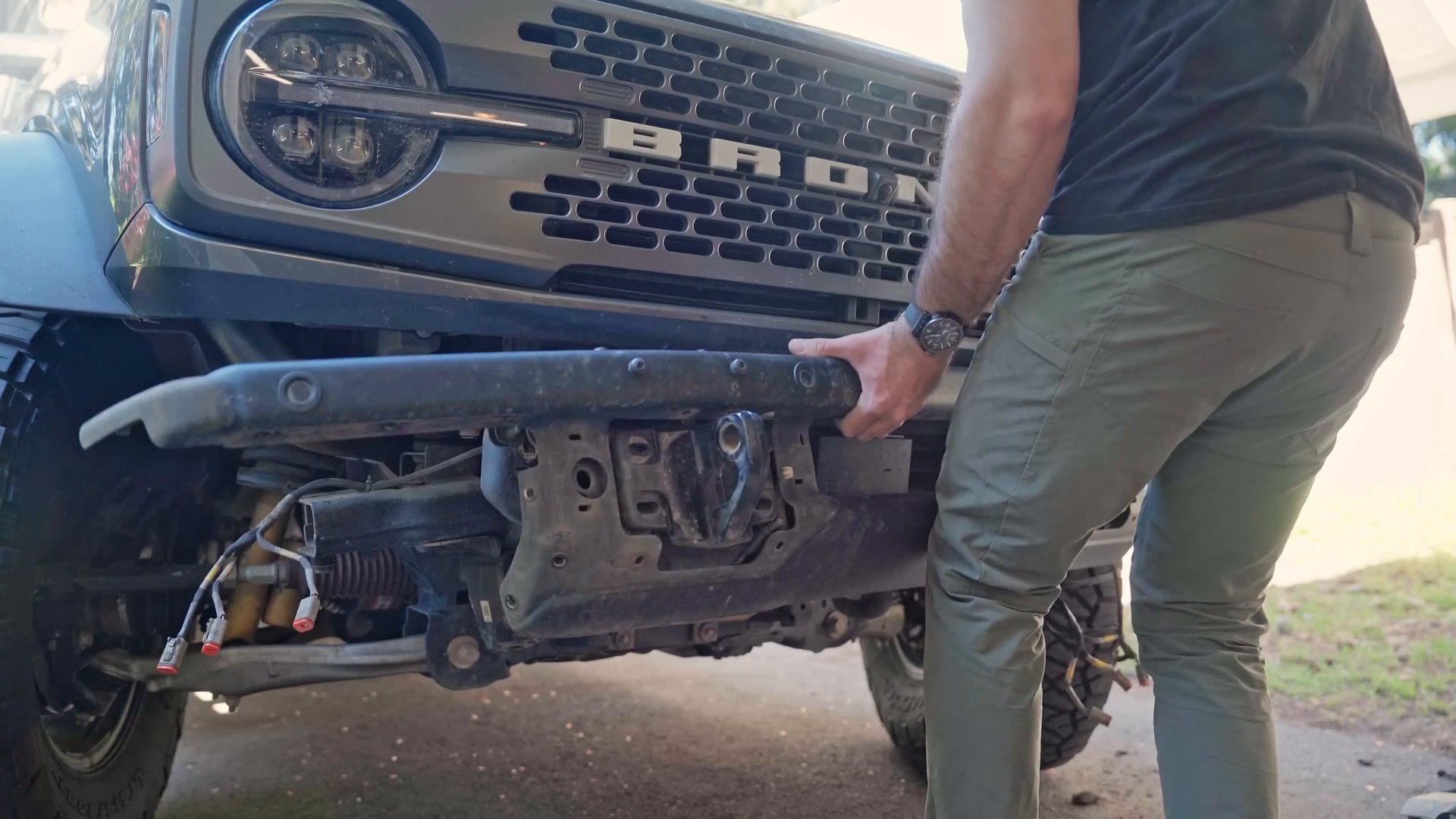 A person wearing green pants and a black shirt is removing or installing a bumper on a truck with large tires and visible suspension components.