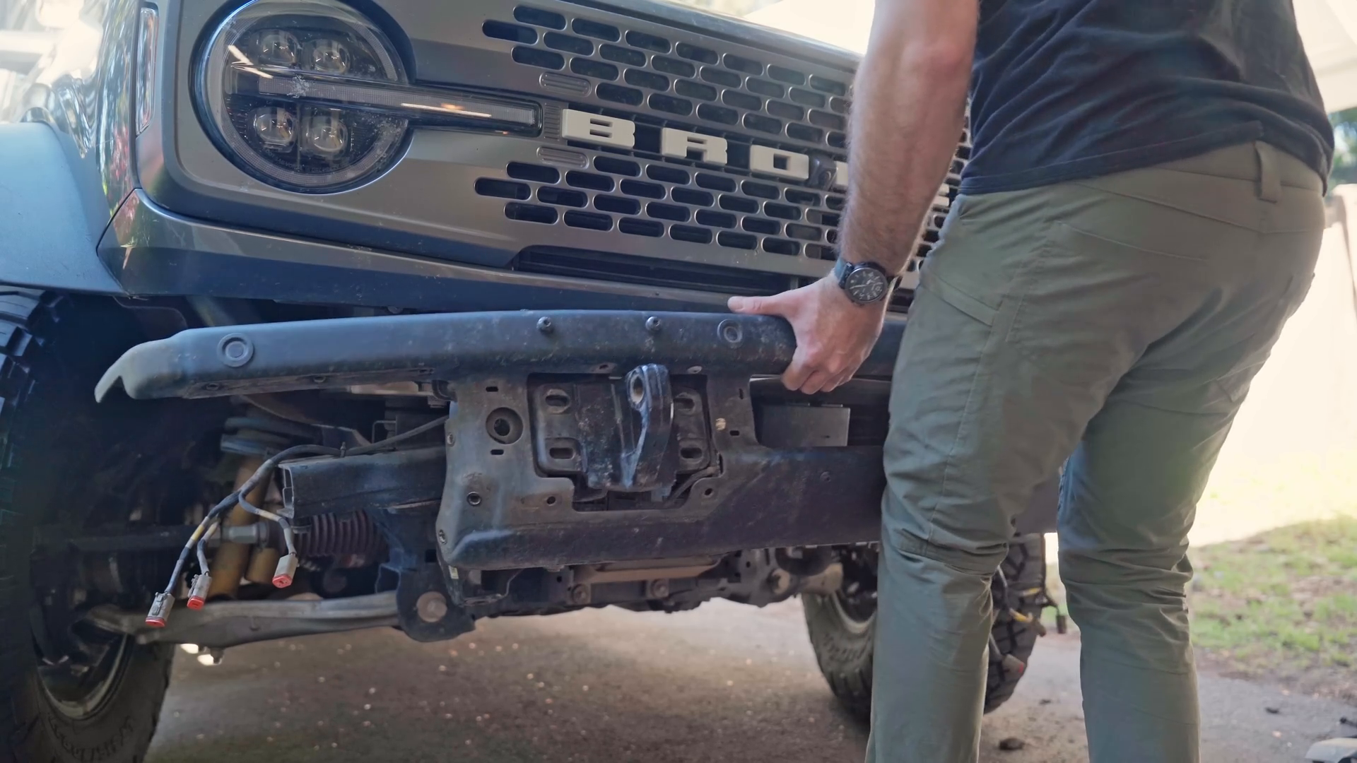 A person is working on the front end of a vehicle, possibly a truck or SUV, handling a detached bumper. The vehicle has prominent round headlights and a grille with lettering. The focus is on the vehicle's chassis and suspension system.