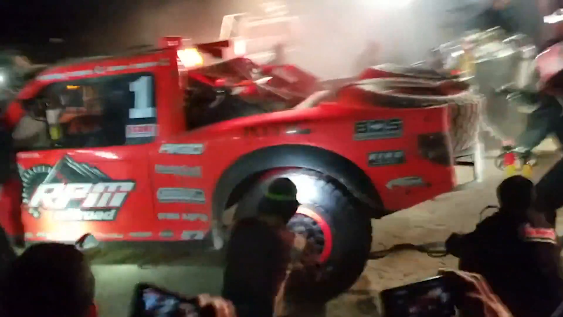 A red off-road race truck with number one on the side is being serviced at night, surrounded by people. Various logos are visible on the truck, including RPM. Bright lights and some spectators with cameras are present.