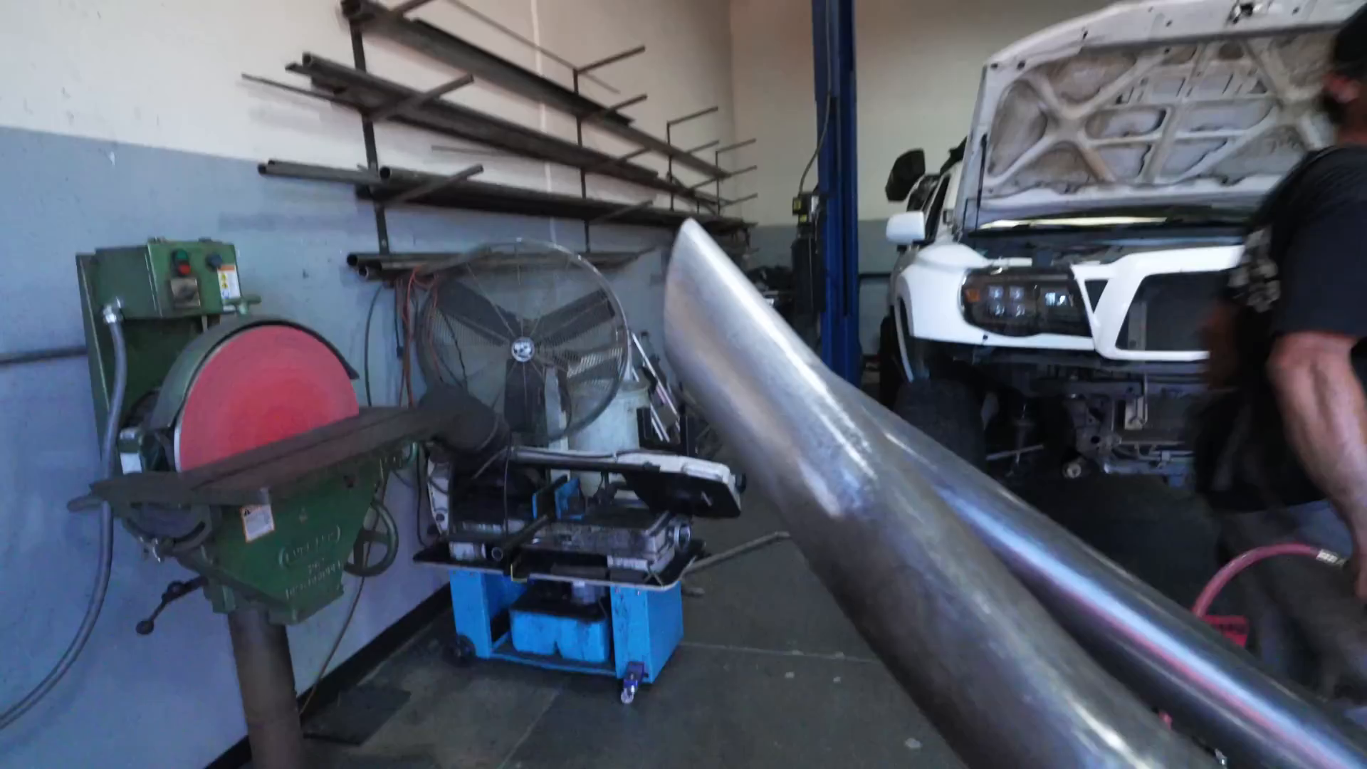 Workshop with metalworking tools and equipment, shelves with metal rods, a large fan, and a white truck with the hood open resembling a prerunner. A person is working near the vehicle.