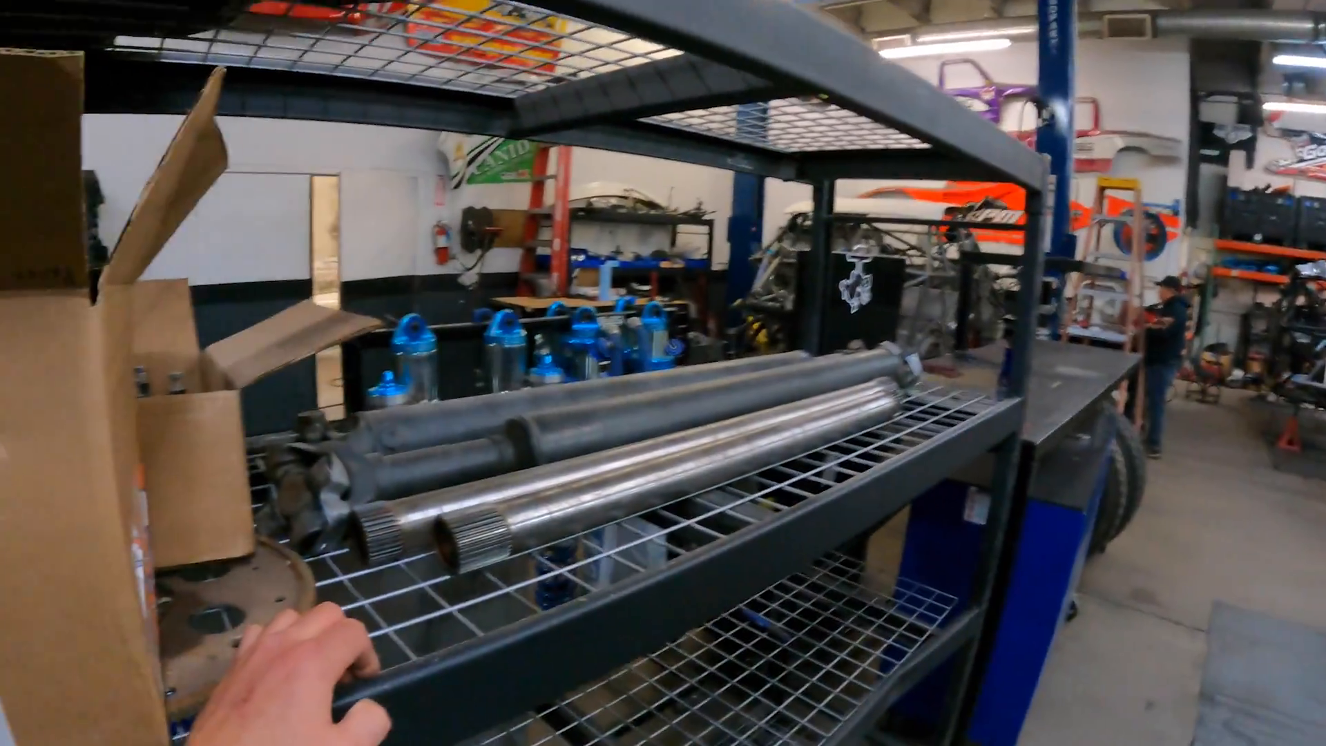 Shelving unit with metal pipes and car parts, blue shock absorbers, a cardboard box, and a workshop area with off-road vehicle frames and equipment.