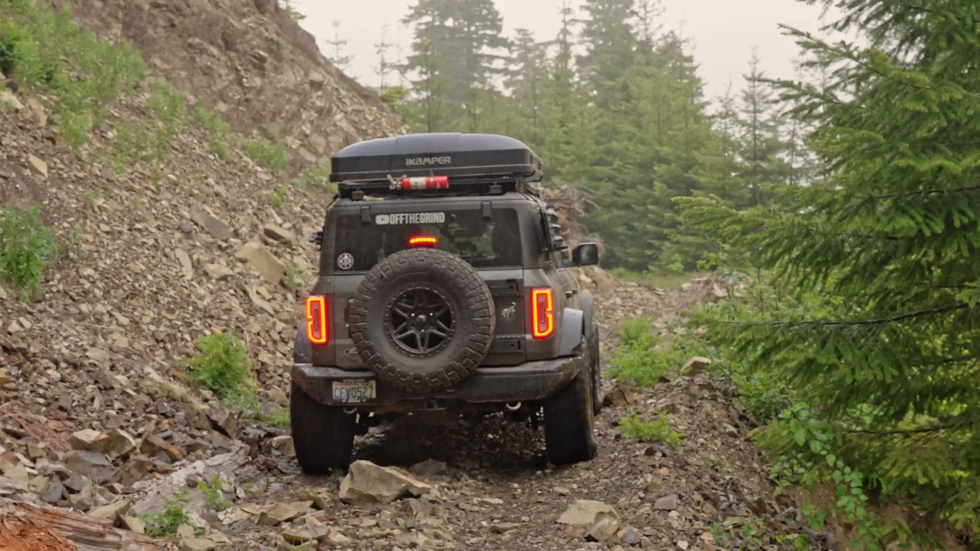 Off-road vehicle with a roof tent and spare tire on a rocky trail surrounded by trees.