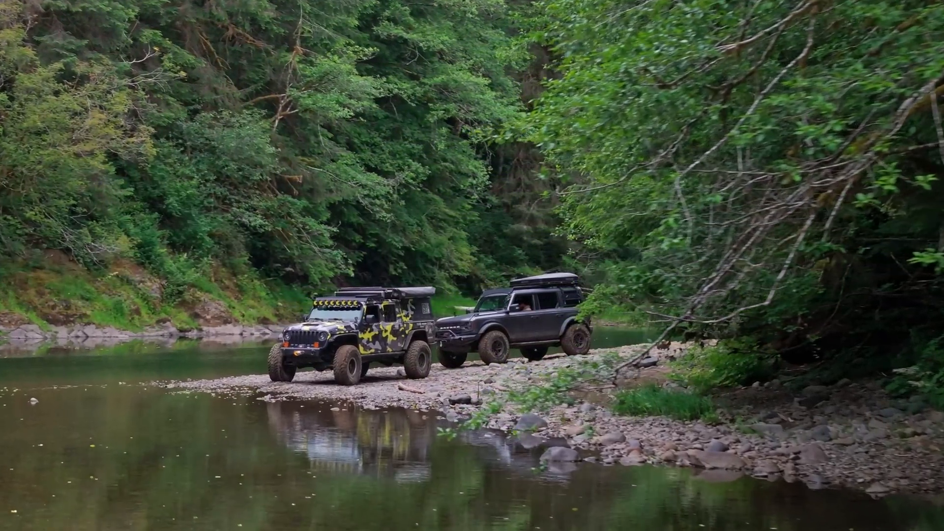 Two off-road vehicles parked on a rocky riverbank, surrounded by dense green forest, one vehicle has a camouflage design and the other is black, bushes and trees overhang the water.