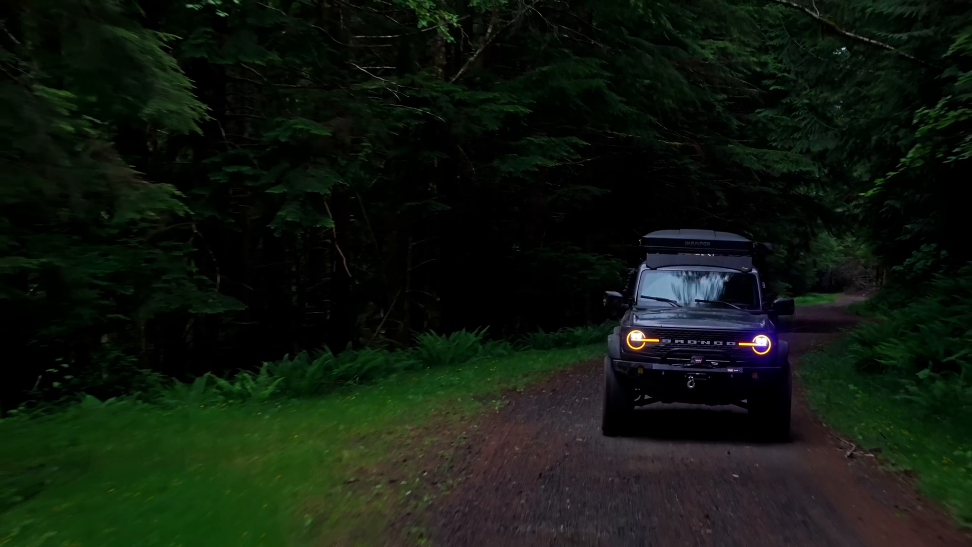 An off-road vehicle on a dirt path surrounded by dense forest, with a roof rack and yellow headlights on.