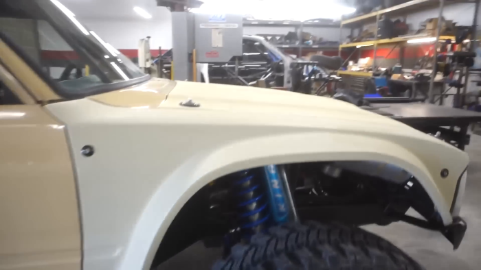 Front left view of a light-colored off-road vehicle with visible suspension and large tire, parked in a workshop with machinery and tools in the background.