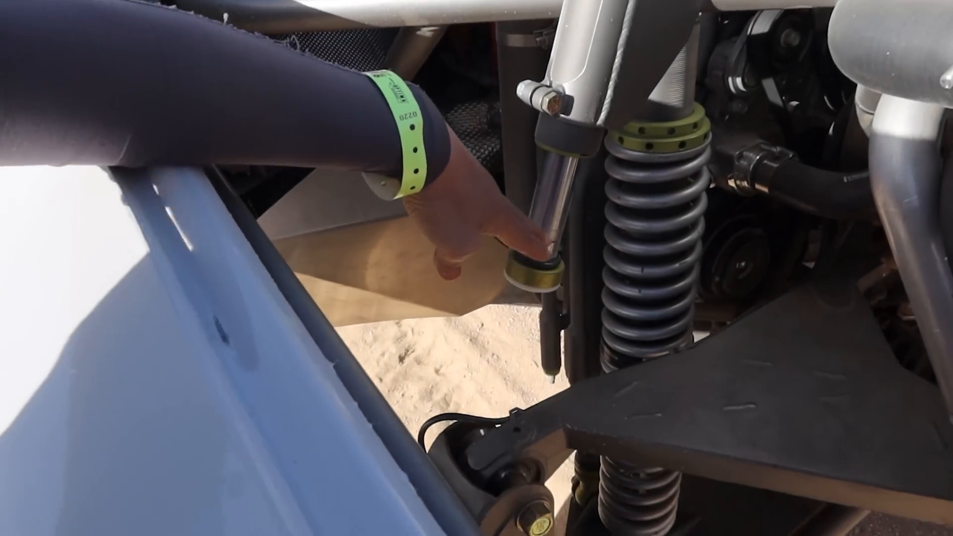 A person with a green wristband points to the suspension system of a prerunner vehicle, highlighting a coilover shock absorber near the vehicle's chassis.