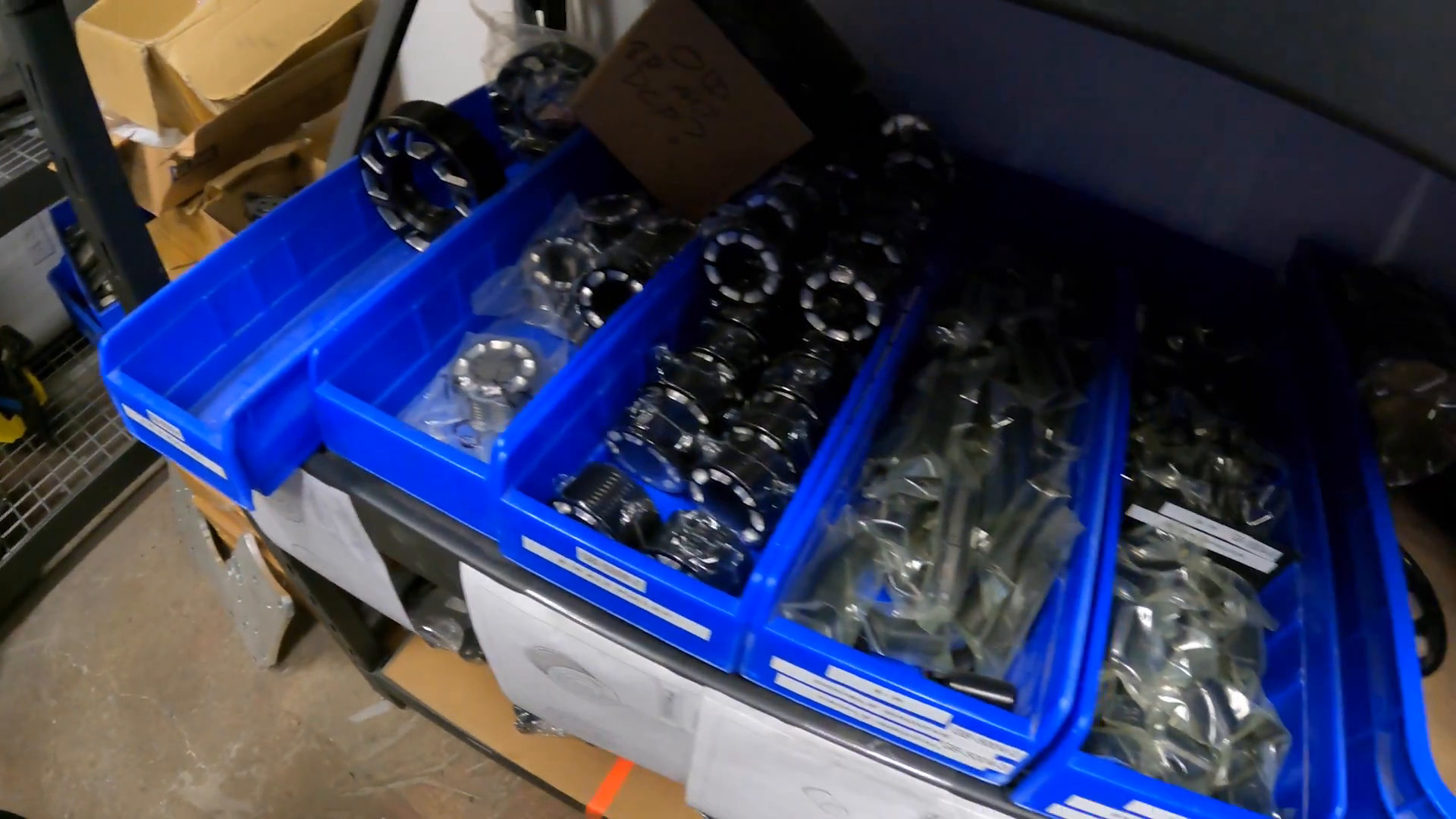 Shelving unit with blue plastic bins containing various metal wheel spacers, labeled for Prerunners, alongside cardboard boxes in a workshop setting.