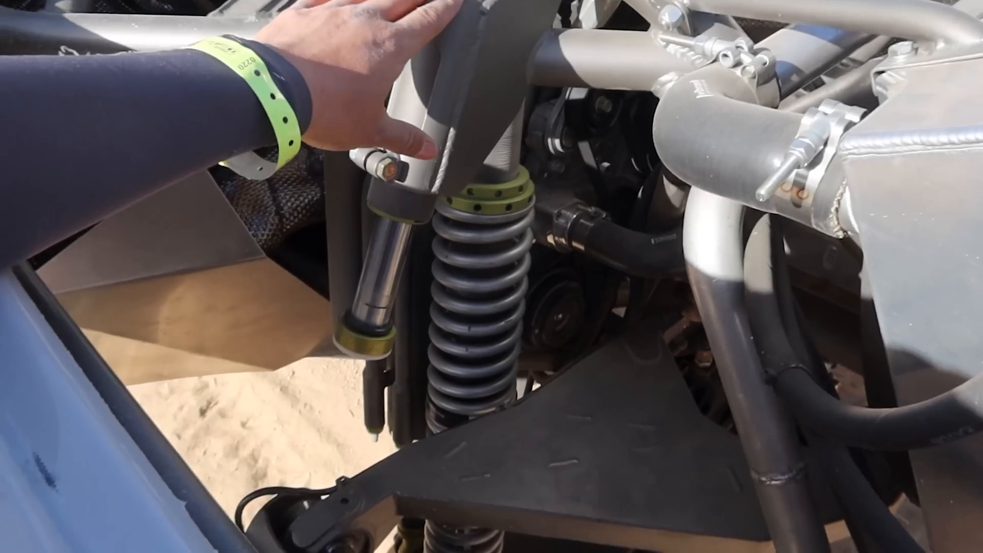 A person wearing a wristband and long-sleeve shirt is examining the suspension system of a prerunner vehicle, focusing on a large coil-over shock absorber with surrounding metal components.