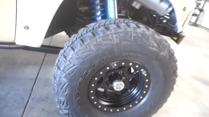 Close-up of a vehicle's all-terrain tire with a black wheel and visible suspension coil in the background, likely part of a prerunner setup.