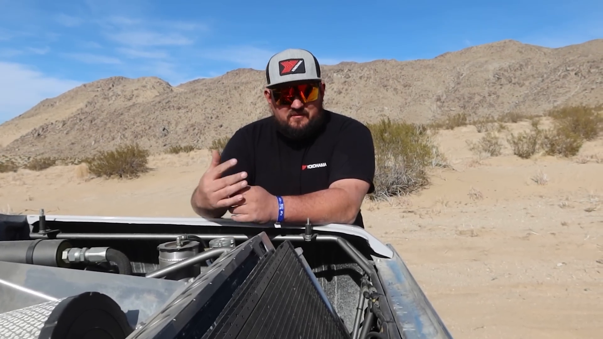 A man wearing sunglasses and a hat stands next to an open area of a Prerunner vehicle with visible components like a radiator and hoses in a desert setting with mountains in the background.