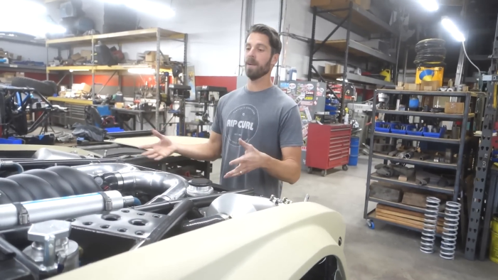 A man in a workshop stands next to the open engine bay of a vehicle, possibly a prerunner truck, with various tools and equipment on shelves and workbenches in the background.