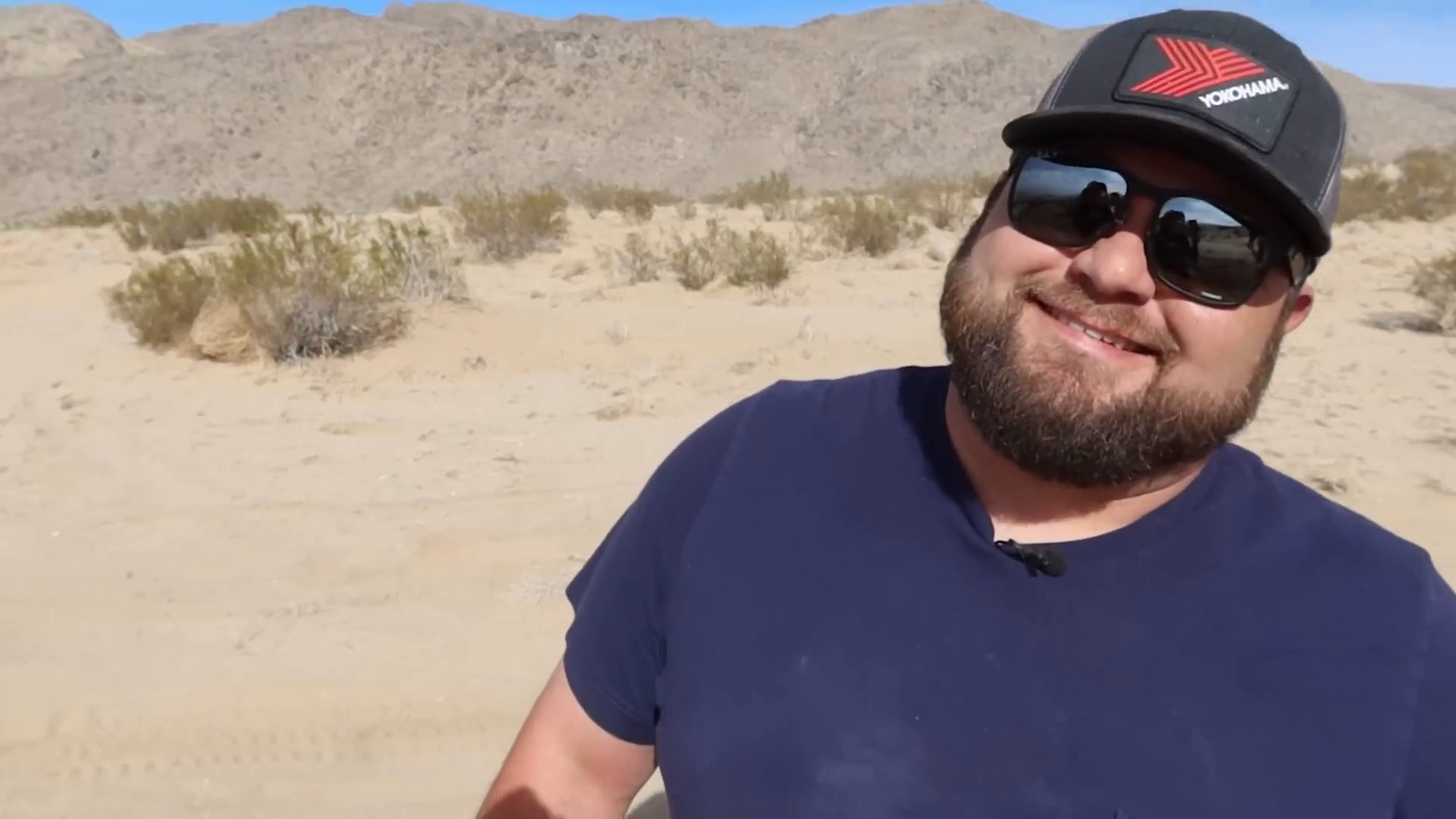 A man wearing a navy blue shirt, sunglasses, and a cap with a Yokohama logo smiling in a desert landscape with sandy terrain and sparse vegetation, mountains in the background.