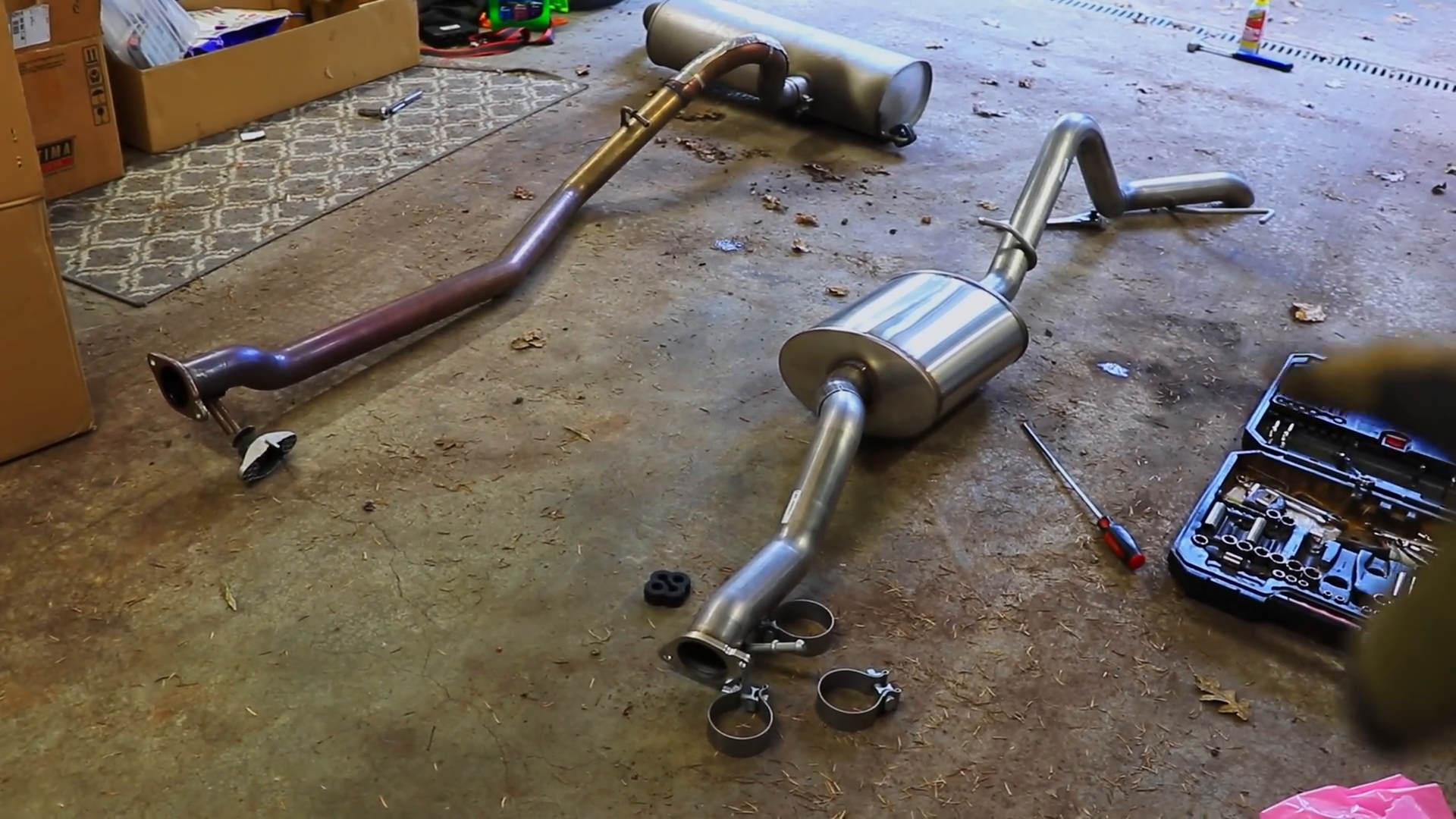 A truck exhaust system with pipes and muffler components on a garage floor, a toolkit, screwdriver, and boxes nearby.