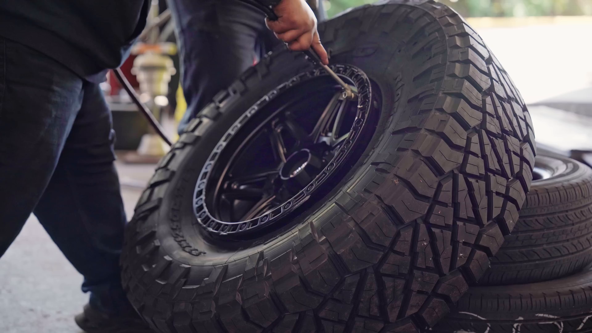 A person installing a black off-road tire onto a rim using a tool in a garage setting.