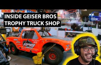 A bright orange trophy truck with RPM Offroad branding in a workshop setting, surrounded by tools and equipment. A man wearing headphones and a camo cap smiling in the foreground. Text overlay reads "Inside Geiser Bros Trophy Truck Shop."